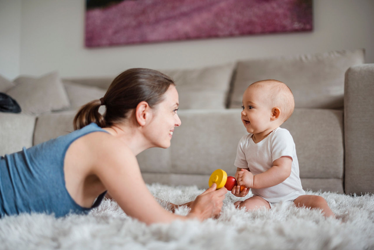 Une femme interagit avec un bebe sur un tapis blanc moelleux dans un salon confortable. Le bebe, vetu d'une grenouillere blanche, tient un jouet colore avec des elements rouges et jaunes. L'arriere-plan presente un canape beige et une �uvre d'art murale floue, creant une atmosphere chaleureuse et accueillante.