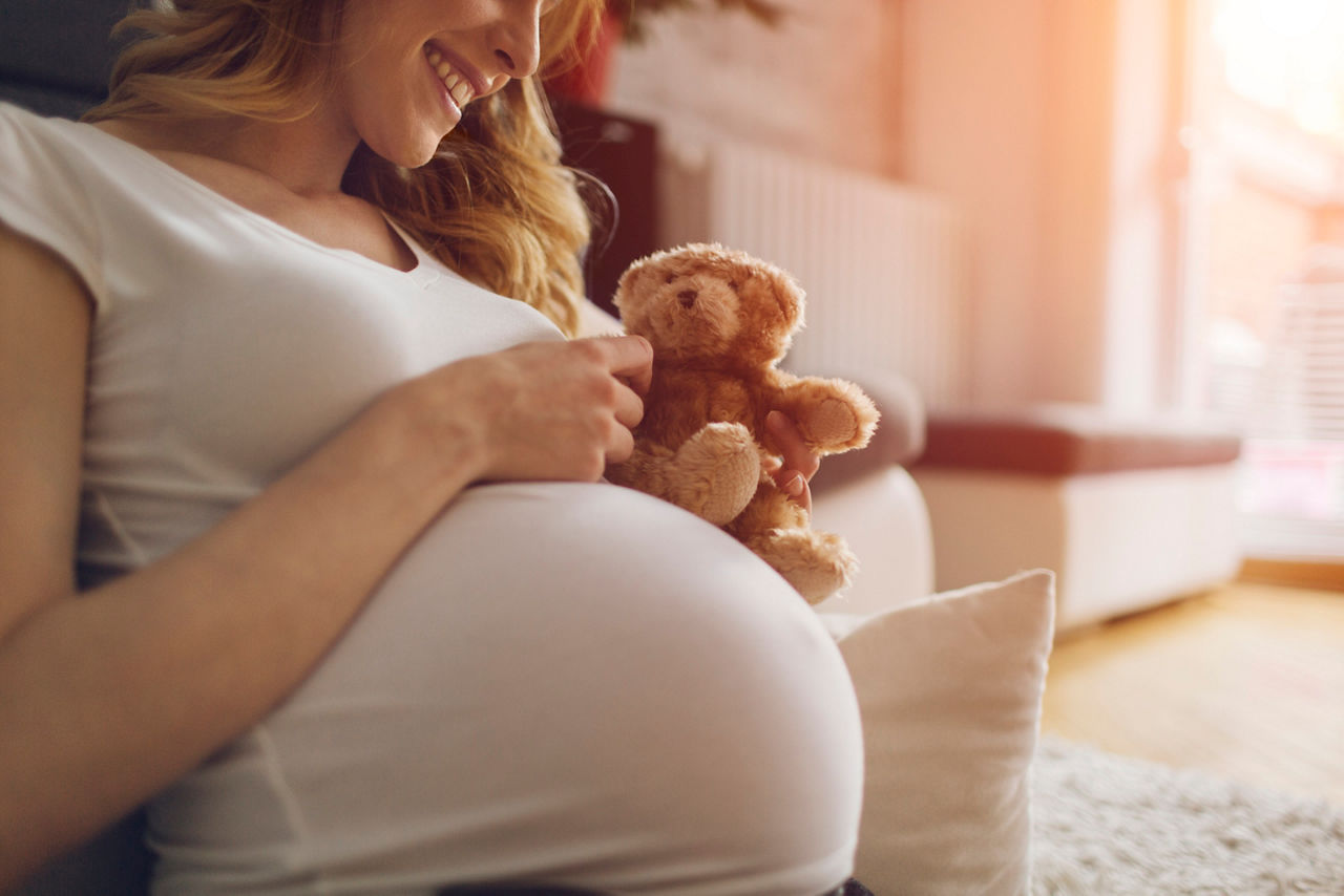 Une femme enceinte souriante est confortablement assise a l'interieur, tenant un petit ours en peluche pres de son ventre rond. Le decor se caracterise par un eclairage chaleureux et un environnement domestique confortable avec des tissus d'ameublement doux. La scene transmet un sentiment d'anticipation et de joie dans la preparation de l'arrivee d'un nouveau bebe.