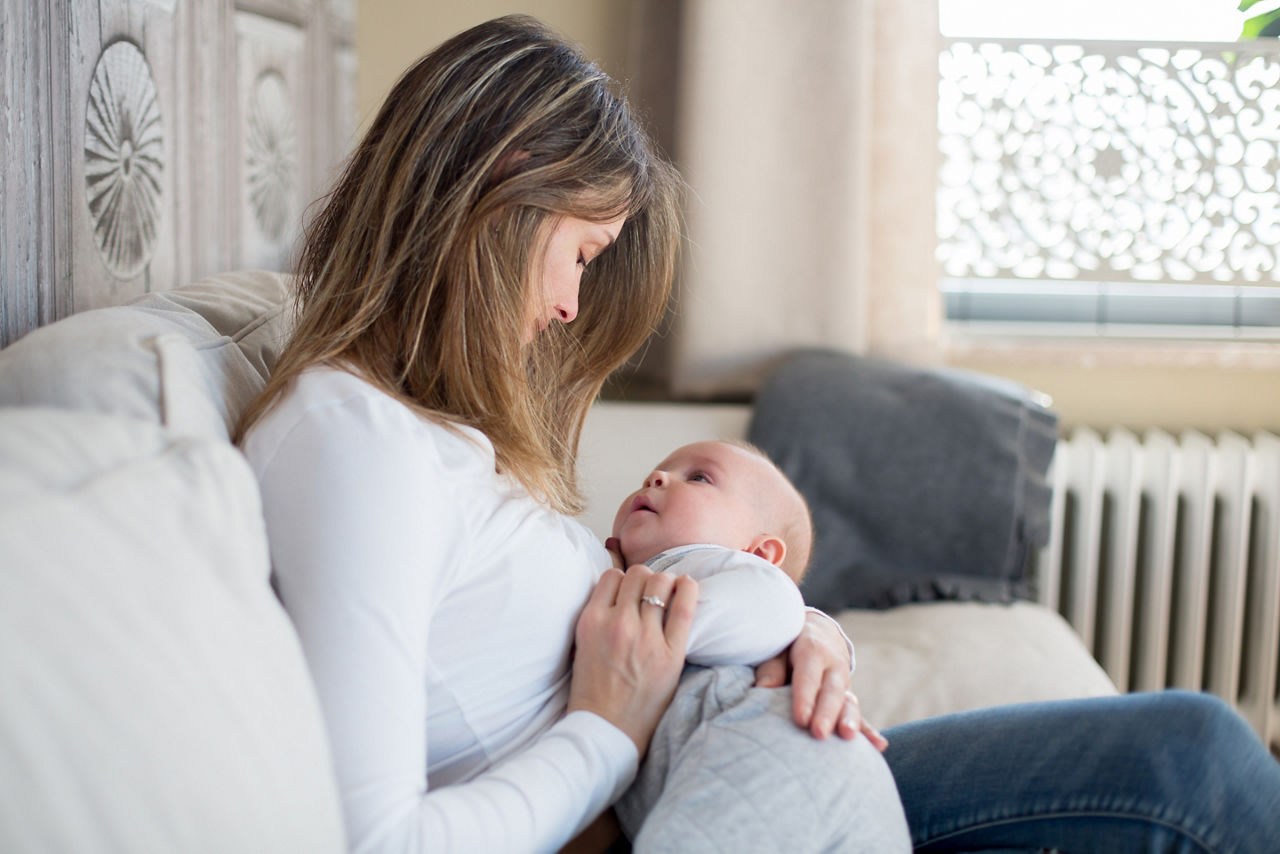 Une femme est assise sur un canape et allaite son bebe dans une piece confortable et faiblement eclairee. Le decor se caracterise par des tons neutres, un panneau decoratif en bois et une fenetre avec des motifs detailles. La scene degage une impression de chaleur et de tendresse dans un environnement familial.