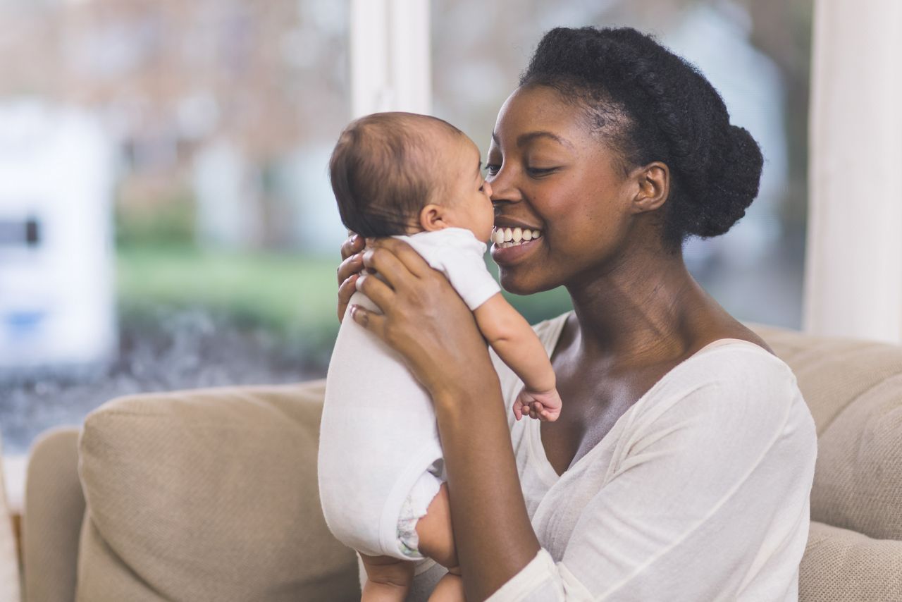 A beautiful young African American mother gently holds her infant daughter up in the air with both hands and kisses her cheek. The baby's eyes are wide open and she looks happy. They are sitting on a couch in their living room.