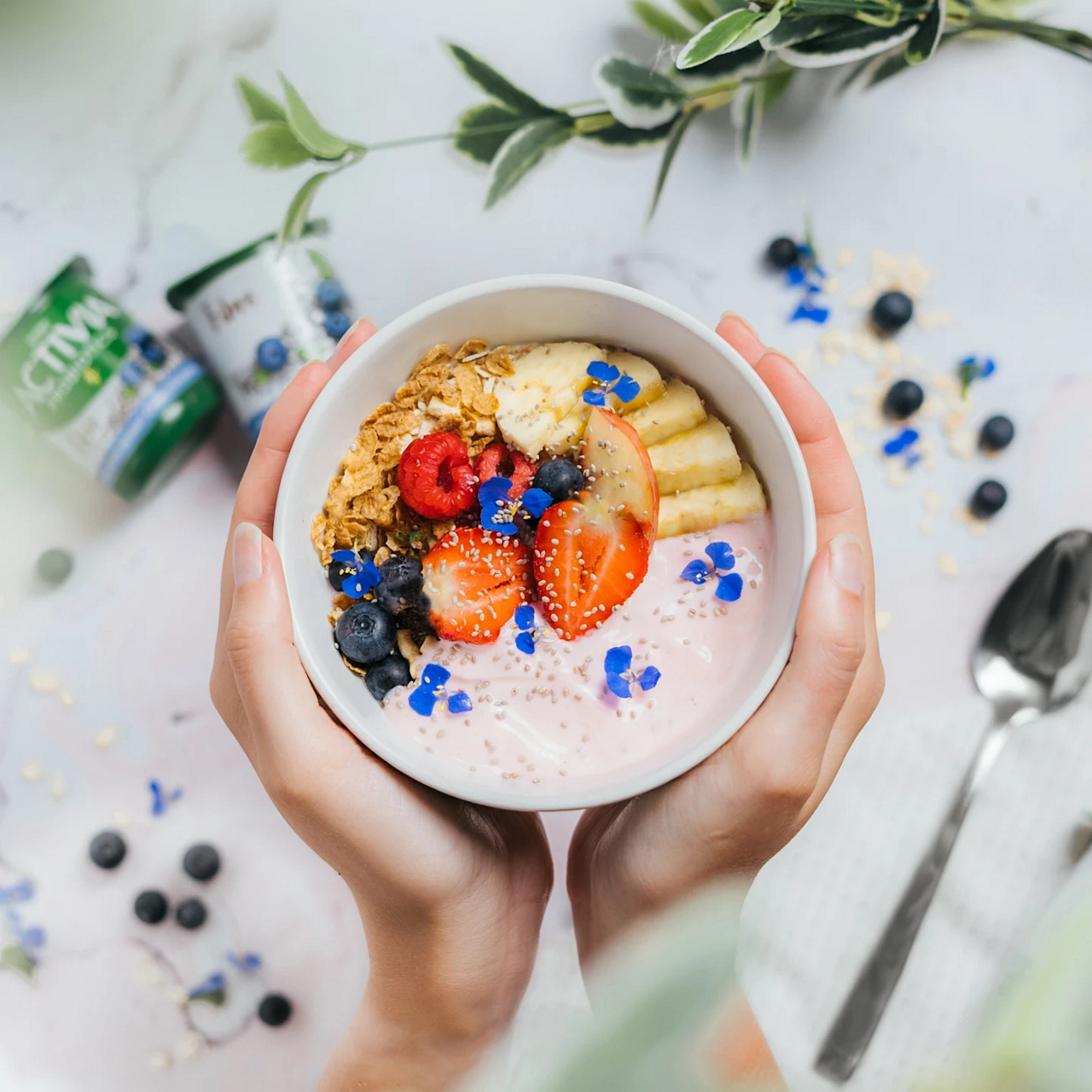 A person holds a white bowl filled with pink yogurt topped with granola, sliced strawberries, banana, blueberries, raspberries, and small blue edible flowers. The bowl is positioned over a light marble surface scattered with oats and blueberries. A spoon and textured napkin sit to the side, while green leaves frame the scene from above. Two small green dairy containers are blurred in the background, suggesting a fresh, healthy breakfast setting.