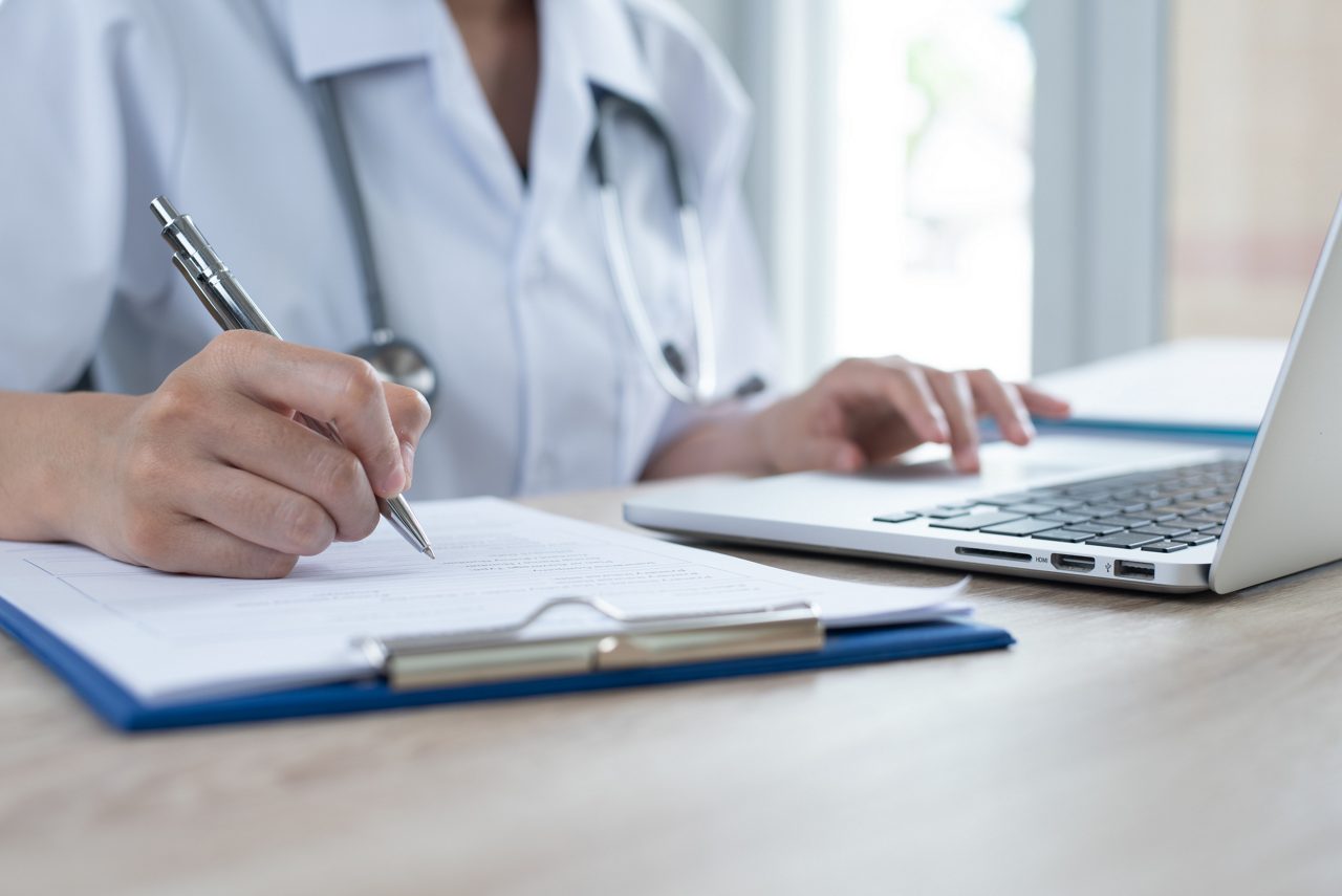 Female doctor working on laptop computer and writing medical insurance from or prescription in doctor office in hospital, closeup.