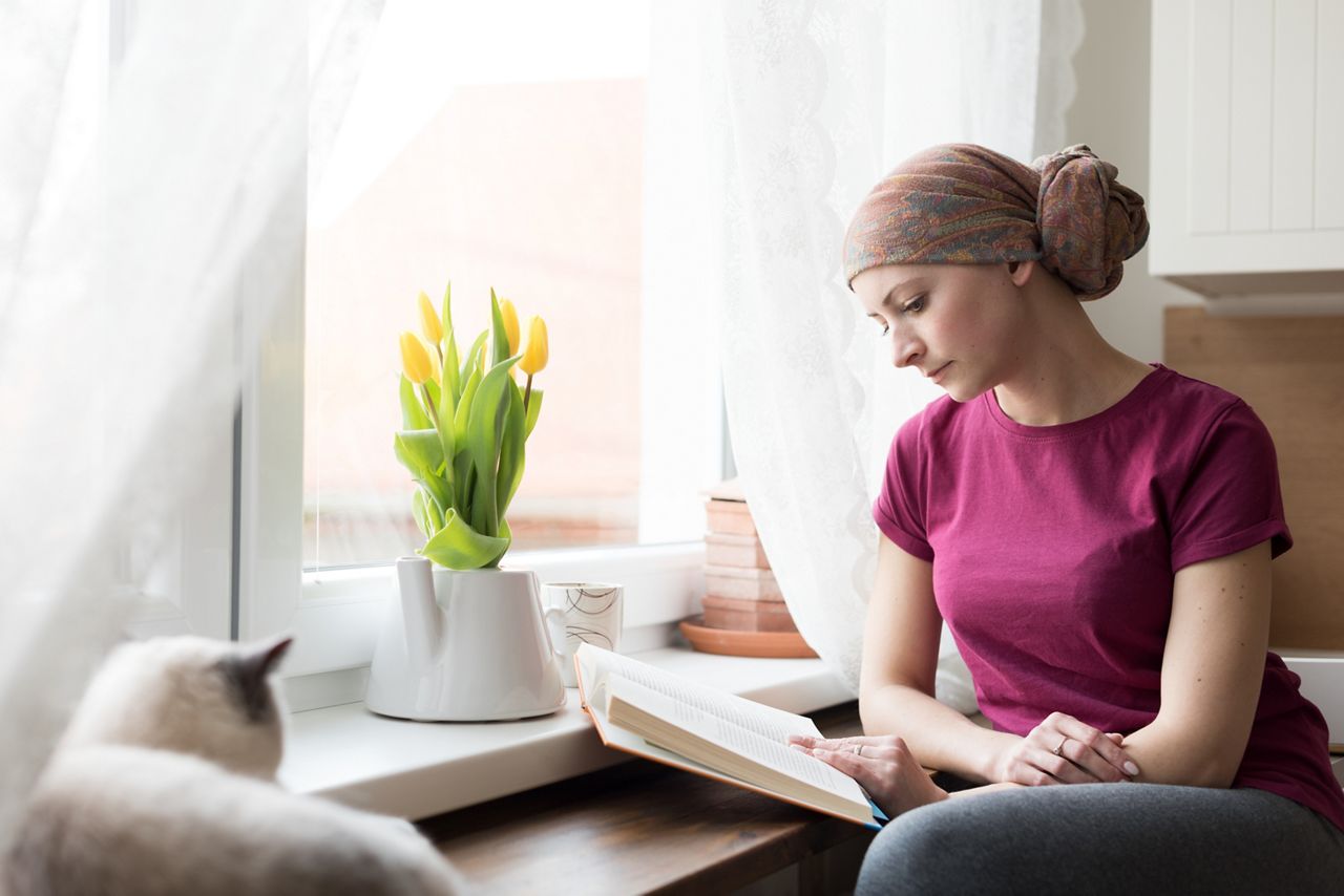 Young adult female cancer patient wearing headscarf sitting in the kitchen with her pet cat, relaxing by reading a book.; Shutterstock ID 1053953330; purchase_order: DNC Thumbnails; job: Webinars 1 (50/189); client: ; other: 