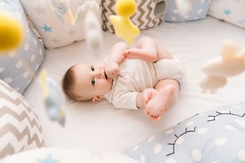 Cute baby lies in a white round bed. Light nursery for young children.  Toys for infant cot. Smiling child playing with mobile of felt and chewing his feet in sunny bedroom.; Shutterstock ID 1068118394; purchase_order: DNC Thumbnails; job: Webinars 1 (50/189); client: ; other: 