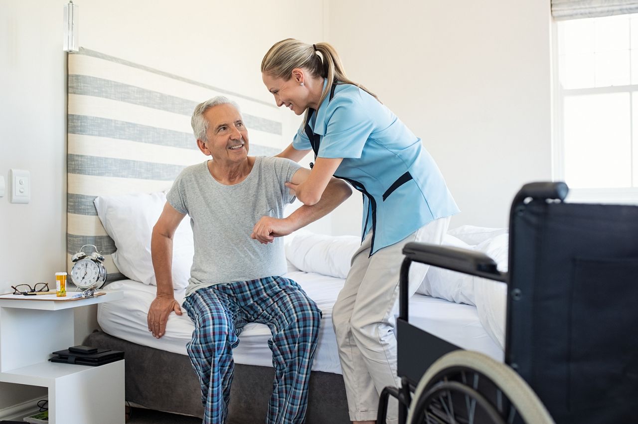 Smiling nurse assisting senior man to get up from bed. Caring nurse supporting patient while getting up from bed and move towards wheelchair at home. Helping elderly disabled man standing up.; Shutterstock ID 1184817247; purchase_order: DNC Thumbnails; job: Videos; client: ; other: 