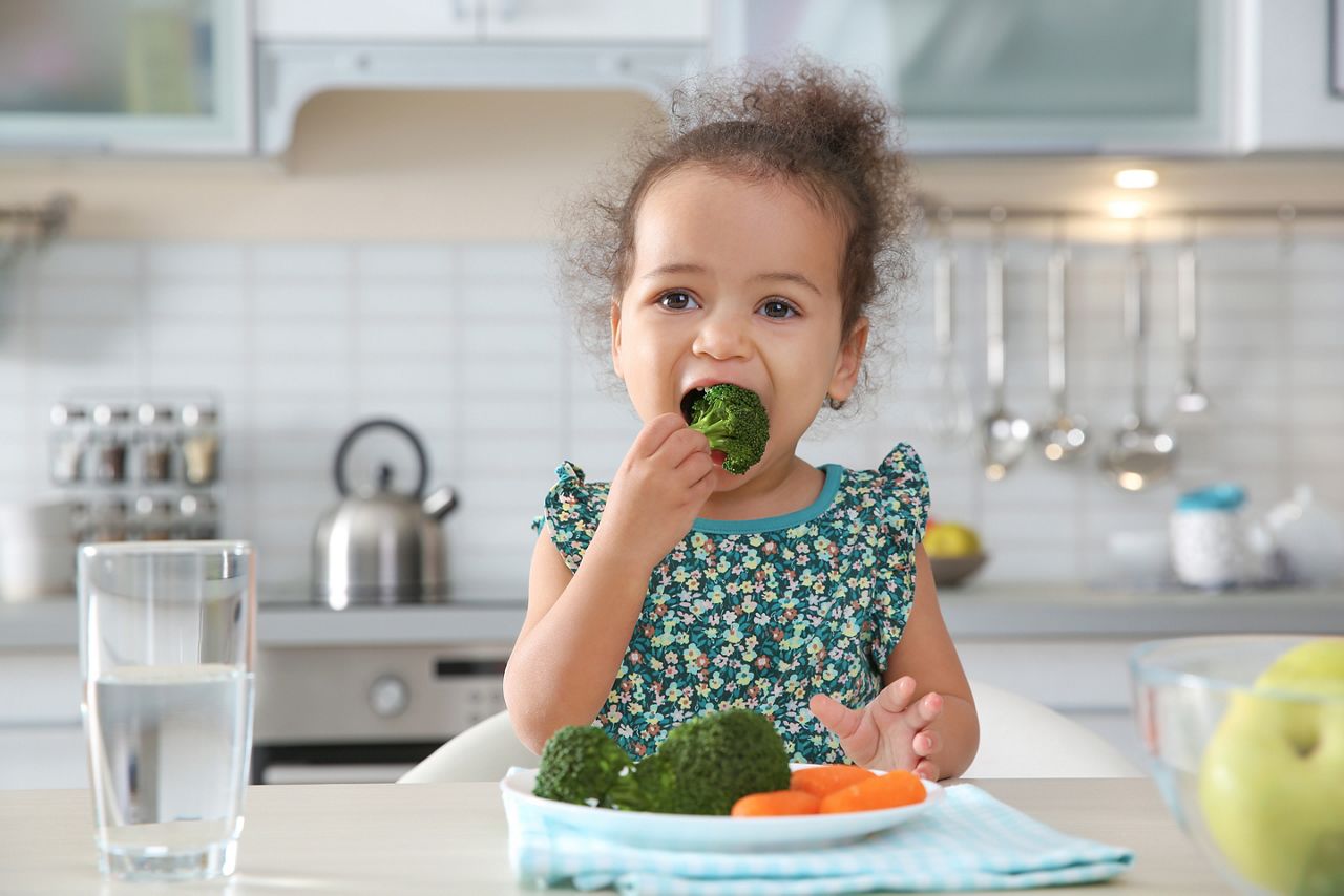 Cute African-American girl eating vegetables at table in kitchen; Shutterstock ID 1293483505; purchase_order: DNC Thumbnails; job: Webinars 2 (50/188); client: ; other: 
