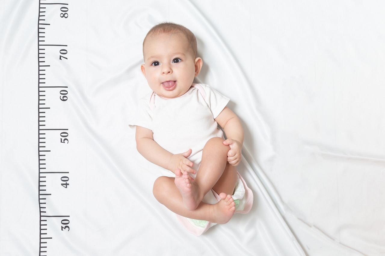 A five month baby in white clothes lying on a bed on which a measuring ruler for growth is drawn. shows the tongue from her mouth and teased; Shutterstock ID 1376349530; purchase_order: DNC Thumbnails; job: Webinars 3 (50/188); client: ; other: 