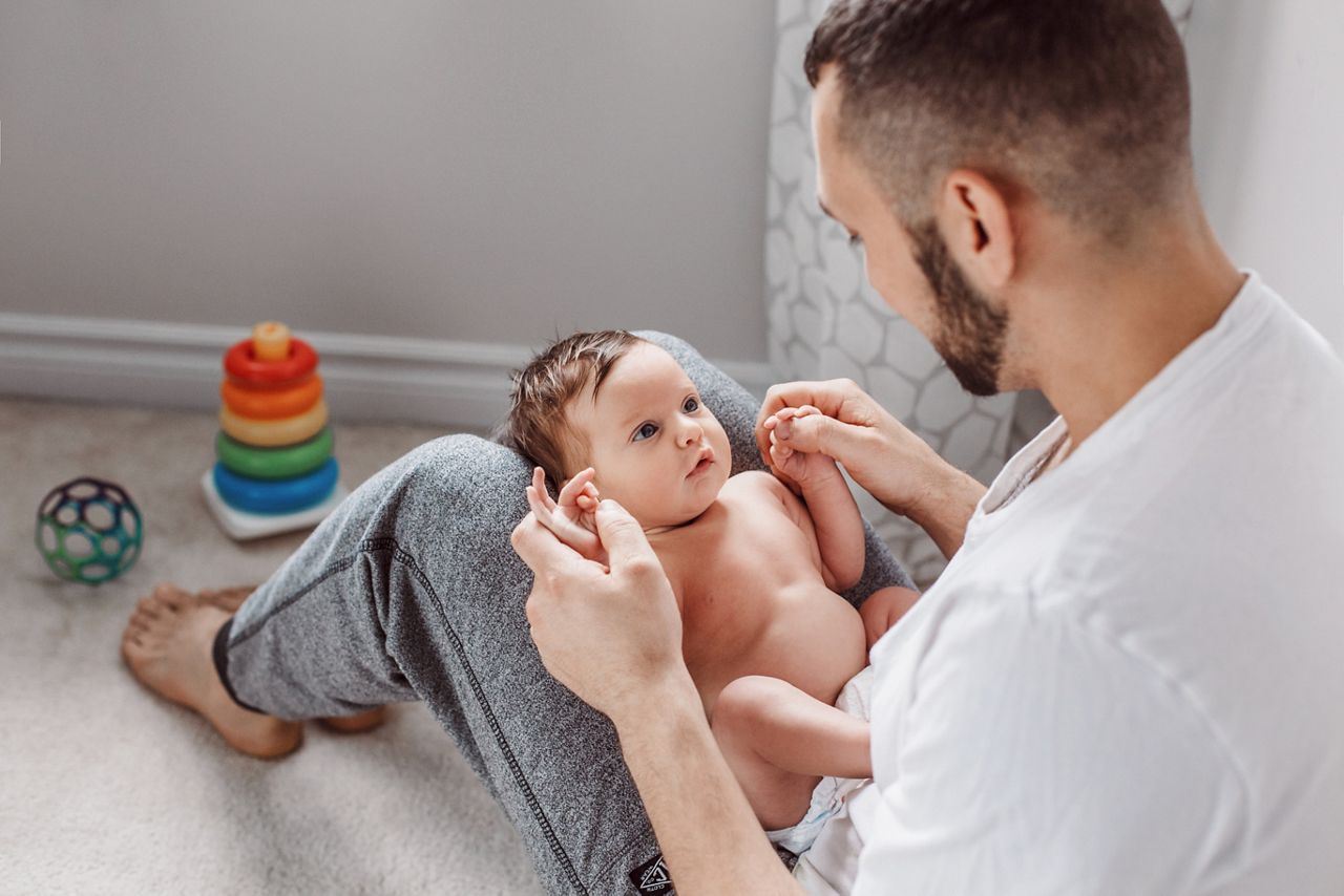 Young Caucasian bearded father holding newborn baby on laps knees. Male man parent rocking talking to child daughter son. Authentic lifestyle documentary moment. Single dad family life.