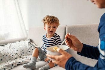 Unhappy child refusing to eat at home with white background while father is trying to deceive the toddler by tricks. Upset child refuse to eat healthy food and demanding bad unhealthy fastfood meals; Shutterstock ID 1549743005; purchase_order: DNC Thumbnails; job: Videos; client: ; other: 