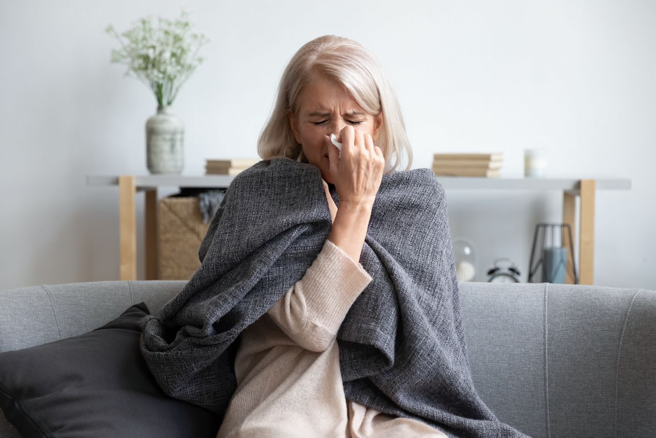 Middle-aged 50s sick frozen woman seated on sofa in living room covered with warm plaid sneezing holding paper napkin blow out runny nose feels unhealthy, seasonal cold, weakened immune system concept; Shutterstock ID 1575687859; purchase_order: DNC Thumbnails; job: Webinars 3 (50/188); client: ; other: 