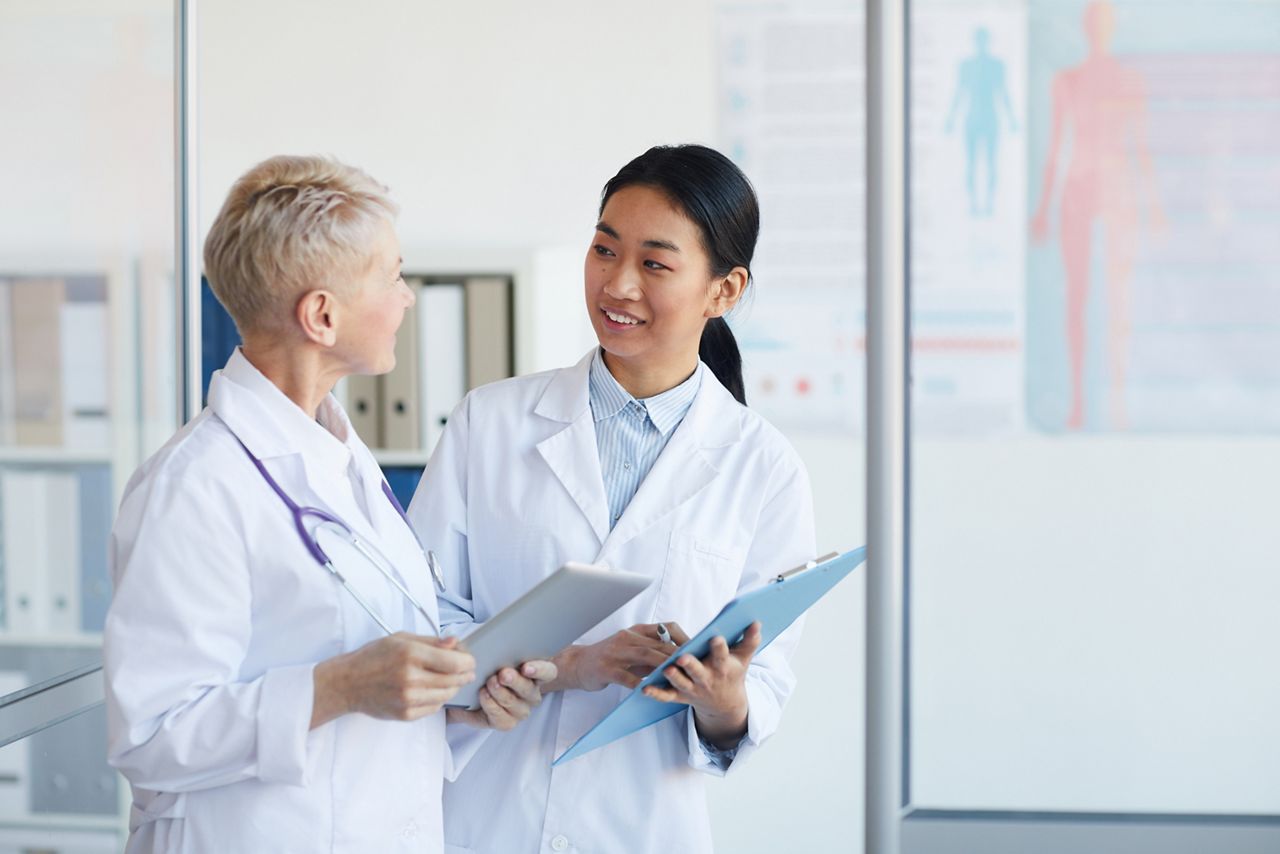 Waist up portrait of young female doctor talking to supervisor and smiling cheerfully while standing in medical office interior, copy space; Shutterstock ID 1674031570; purchase_order: DNC Thumbnails; job: Collections; client: ; other: 