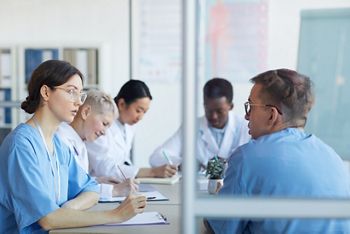 Portrait of young female doctor talking to colleague while sitting at table during medical council or conference in clinic, copy space; Shutterstock ID 1674031651; purchase_order: DNC Thumbnails; job: Webinars 3 (50/188); client: ; other: 