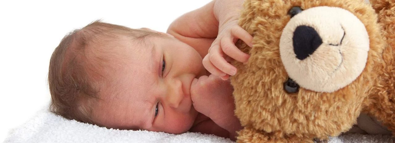 Ein Neugeborenes liegt auf einer weißen Decke und hält einen braunen Teddybären eng an sich.