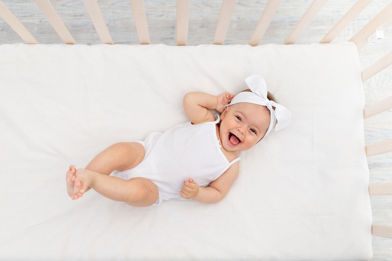baby girl 6 months old lies in a crib in the nursery with white clothes on her back and laughs, looks at the camera, baby's morning, baby products concept; Shutterstock ID 1780775009; purchase_order: DNC Thumbnails; job: Webinars 2 (50/188); client: ; other: 