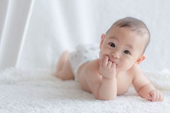 Asian baby boy lying on his tummy on a piece of white fluffy cloth chewing his hands and smiling at the camera; Shutterstock ID 1786349282; purchase_order: SN event thumbnails; job: ; client: ; other: 