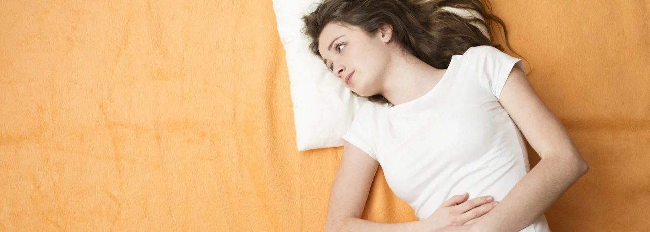 Eine junge Frau liegt mit schmerzverzerrtem Gesicht und den Händen auf dem Bauch auf einem orangefarbenen Bett.