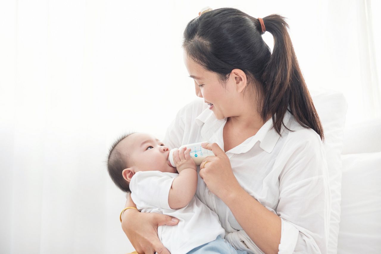 Portrait of a happy asian mother giving bottle feeding to her baby in white bedroom.  Baby is drinking milk from a bottle hold by the mother.; Shutterstock ID 1847377969; purchase_order: DNC Thumbnails; job: Publications; client: ; other: Replace Fishwife
