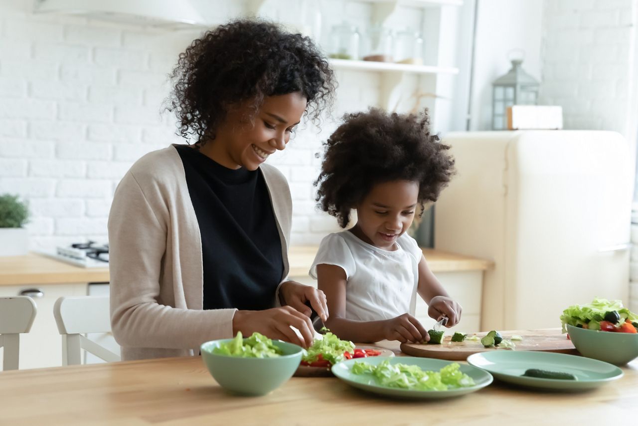Smiling African American mother with daughter cooking salad together, chopping fresh vegetables on board, sitting at wooden kitchen table, caring mum and child preparing meal, having fun; Shutterstock ID 1854376573; purchase_order: 25 thumbnail photos ; job: ; client: ; other: 