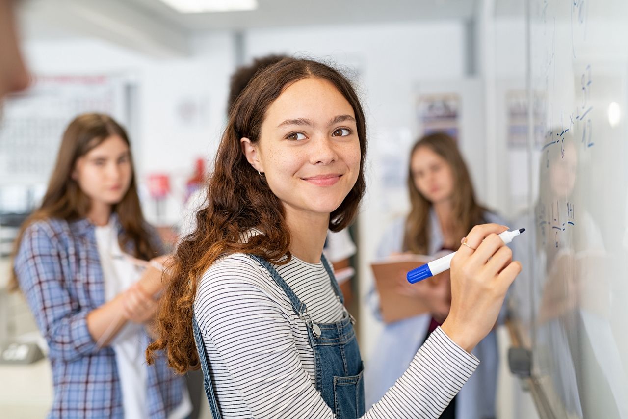 Portrait of smiling girl solving math equation on white board. College student thinking and solving arithmetic sum with classmates. Smart young woman writing on white board during class.; Shutterstock ID 1904756506; purchase_order: DNC Thumbnails; job: Webinars 1 (50/189); client: ; other: 