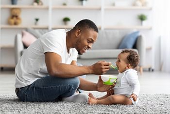 Father's Care. Loving Black Dad Feeding His Cute Baby Son From Spoon At Home, Young African American Daddy Giving Healthy Food To His Little Toddler Child While They Relaxing Together In Living Room; Shutterstock ID 2022074738; purchase_order: 25 thumbnail photos ; job: ; client: ; other: 