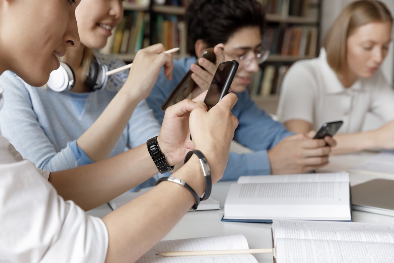 Group of young gen Z students browsing Internet in campus library, using smartphones at work table with books, collaborating on project for class, chatting online. Close up of hands holding phone; Shutterstock ID 2111420600; purchase_order: DNC Thumbnails; job: Webinars 1 (50/189); client: ; other: 