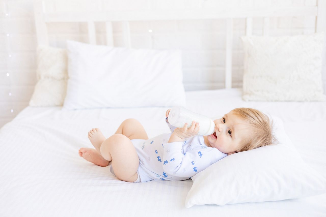 cute baby with a bottle of milk sucks holding it in his handslying on her back on a baby bed in a children's bright room, baby food concept; Shutterstock ID 2148870983; purchase_order: DNC thumbnails ; job: Allergy papers; client: ; other: 