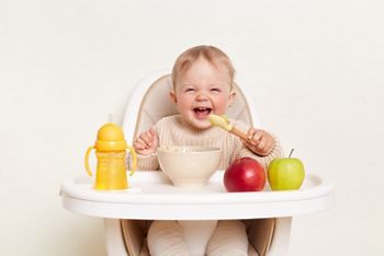 Baby wearing knitted sweater sitting in high chair and feels hungry, holding spoon and eating puree or porridge, enjoying apples and water in yellow bottle, posing isolated on white background.; Shutterstock ID 2169821577; purchase_order: 25 thumbnail photos ; job: ; client: ; other: 