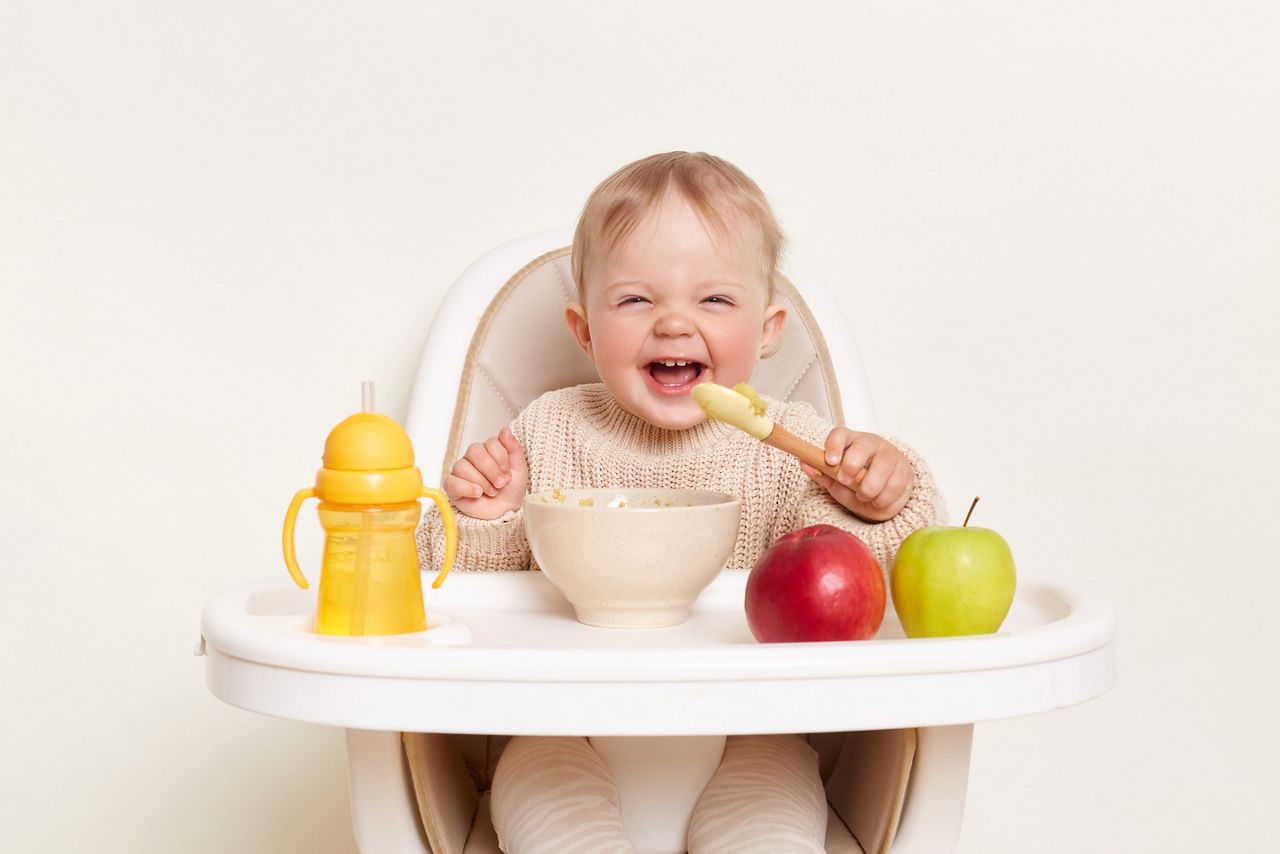 Baby wearing knitted sweater sitting in high chair and feels hungry, holding spoon and eating puree or porridge, enjoying apples and water in yellow bottle, posing isolated on white background.; Shutterstock ID 2169821577; purchase_order: 25 thumbnail photos ; job: ; client: ; other: 