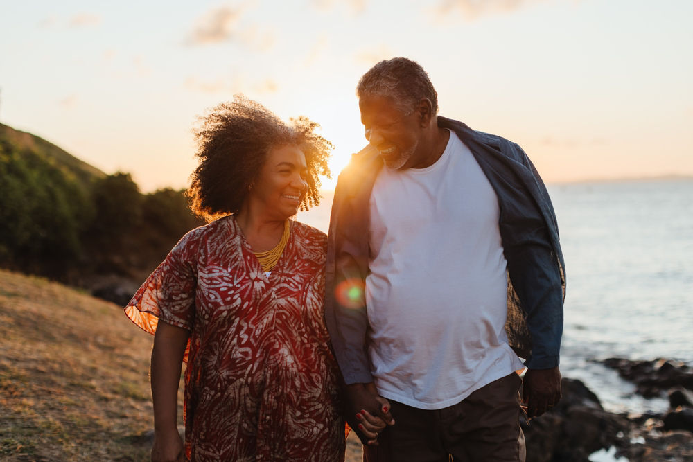 An older couple walking hand-in-hand by the water during a sunset, radiating feelings of romance, joy, and togetherness. Demonstrating elderly companionship.