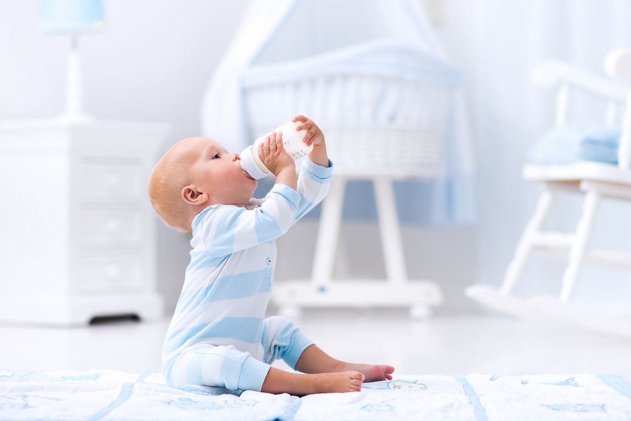 Adorable baby boy playing on a blue floor mat and drinking milk from a bottle in a white sunny nursery with rocking chair and bassinet. Bedroom interior with infant crib. Formula drink for infant.; Shutterstock ID 481192738; purchase_order: DNC thumbnails ; job: Allergy papers; client: ; other: 
