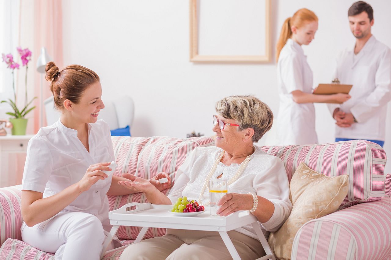 Elderly lady sitting on a couch with a tray and a young nurse giving her medicines in private clinic; Shutterstock ID 530192146; purchase_order: DNC Thumbnails; job: Publications; client: ; other: Replace Fishwife