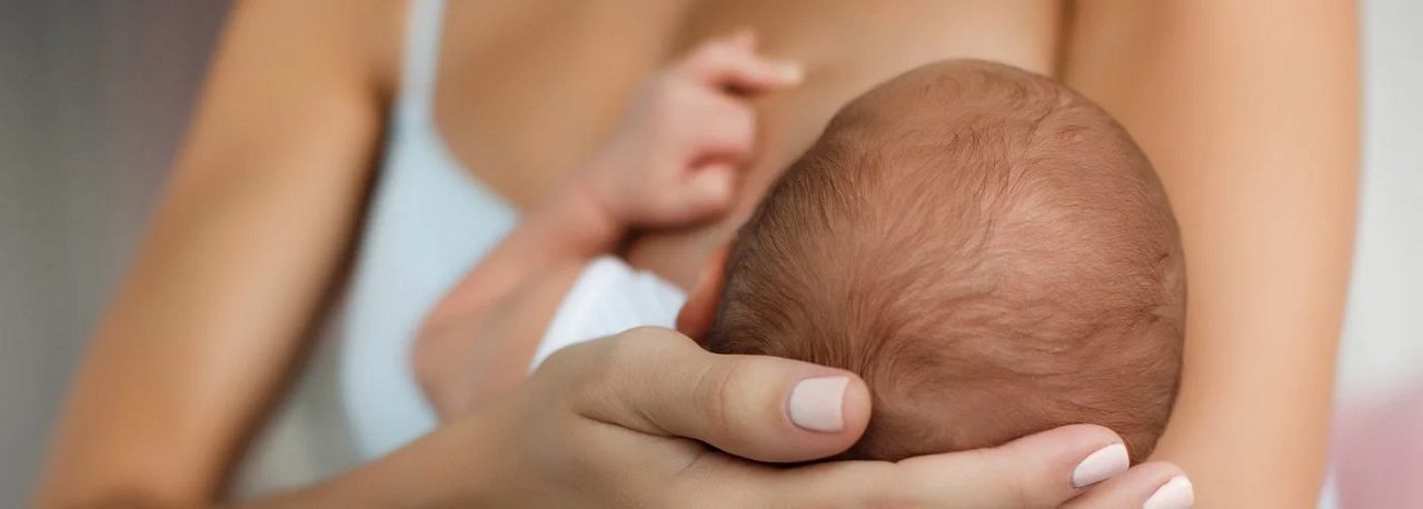 Eine Frau hält ein Baby und stillt es an der Brust, der Kopf des Babys wird mit einer Hand gestützt.
