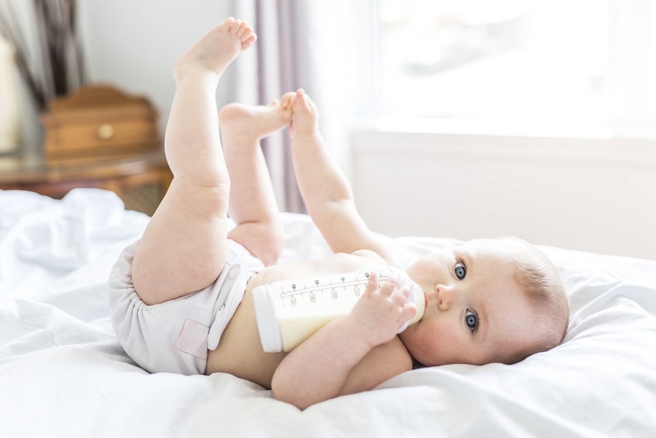 A Pretty baby girl drinks water from bottle lying on bed. Child weared diaper in nursery room.; Shutterstock ID 627983705; purchase_order: DNC Thumbnails; job: Webinars 2 (50/188); client: ; other: 