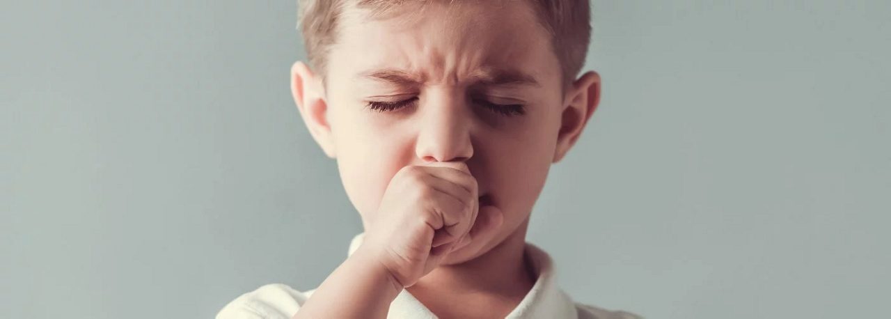 Ein Junge hält seine Faust vor den Mund und hustet, im Hintergrund ist eine einfarbige Wand zu sehen.