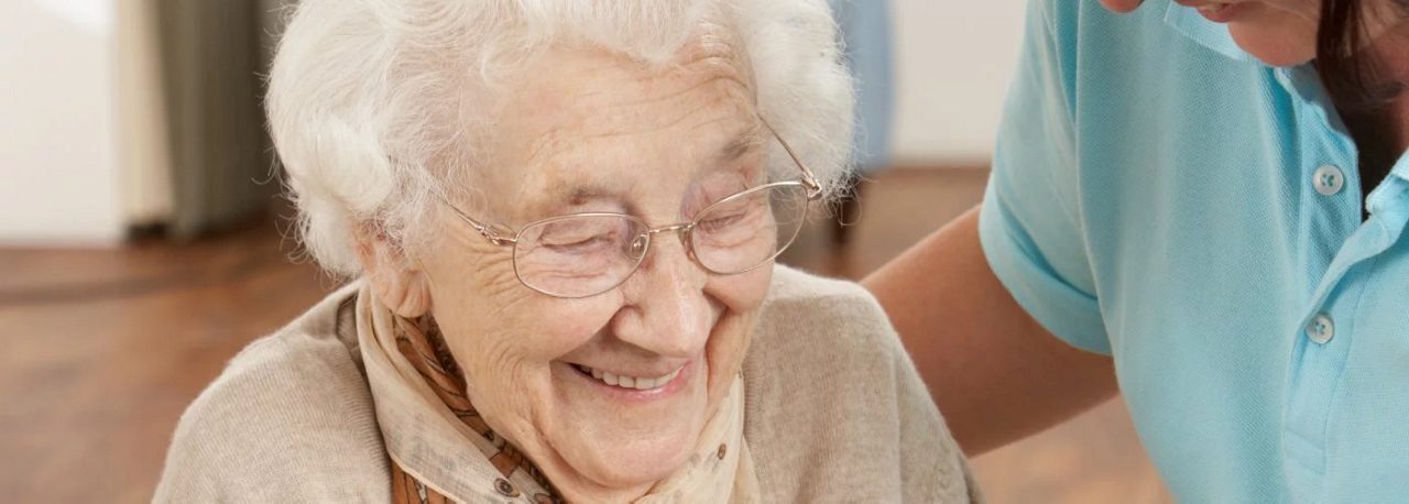 Eine ältere Frau mit Brille lächelt, während eine Pflegekraft in blauer Kleidung sie fürsorglich am Arm hält.