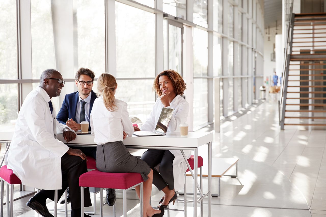 Four doctors talking at a table in a modern hospital lobby; Shutterstock ID 737503960; purchase_order: DNC Thumbnails; job: Webinars 1 (50/189); client: ; other: 