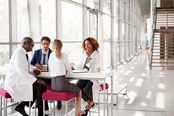 Four doctors talking at a table in a modern hospital lobby; Shutterstock ID 737503960; purchase_order: DNC Thumbnails; job: Webinars 1 (50/189); client: ; other: 