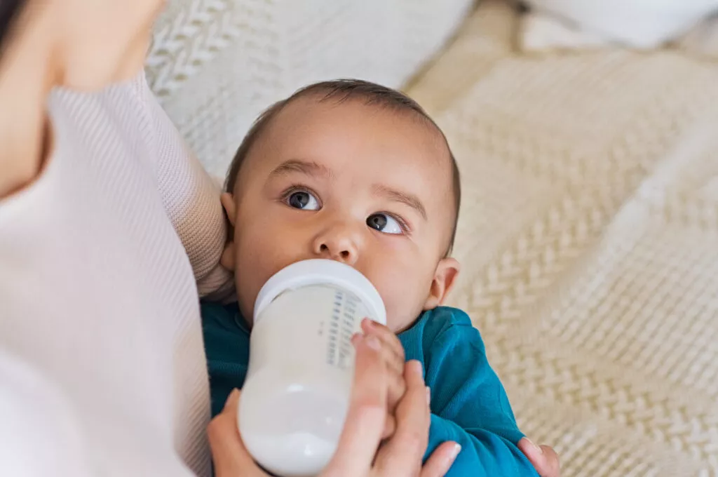 Cute baby drinking milk from baby bottle while looking his mom. Mother feeding son infant from bottle. Little boy drinking milk from bottle at home.