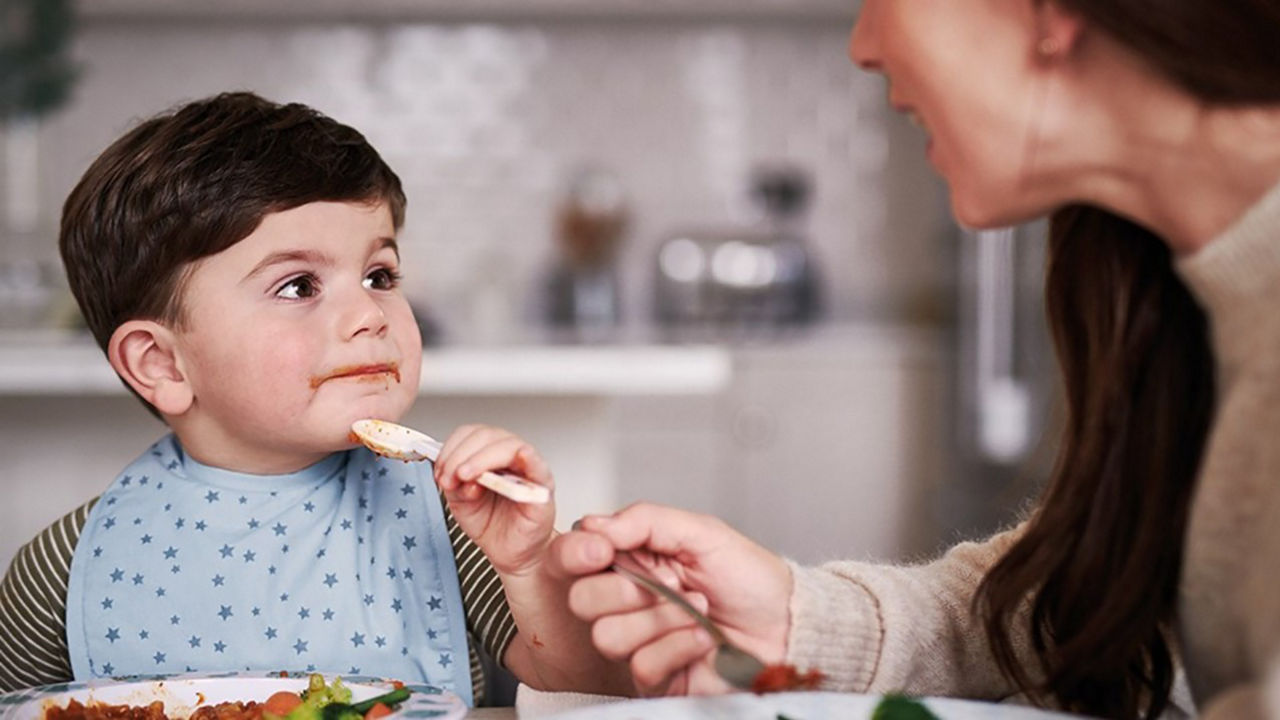 Niño pequeño comiendo con cuchara en la mesa junto a su madre, representando hábitos saludables de alimentación y cuidado del bebé, contenido de Nutricia Club (Cuidado bebé)