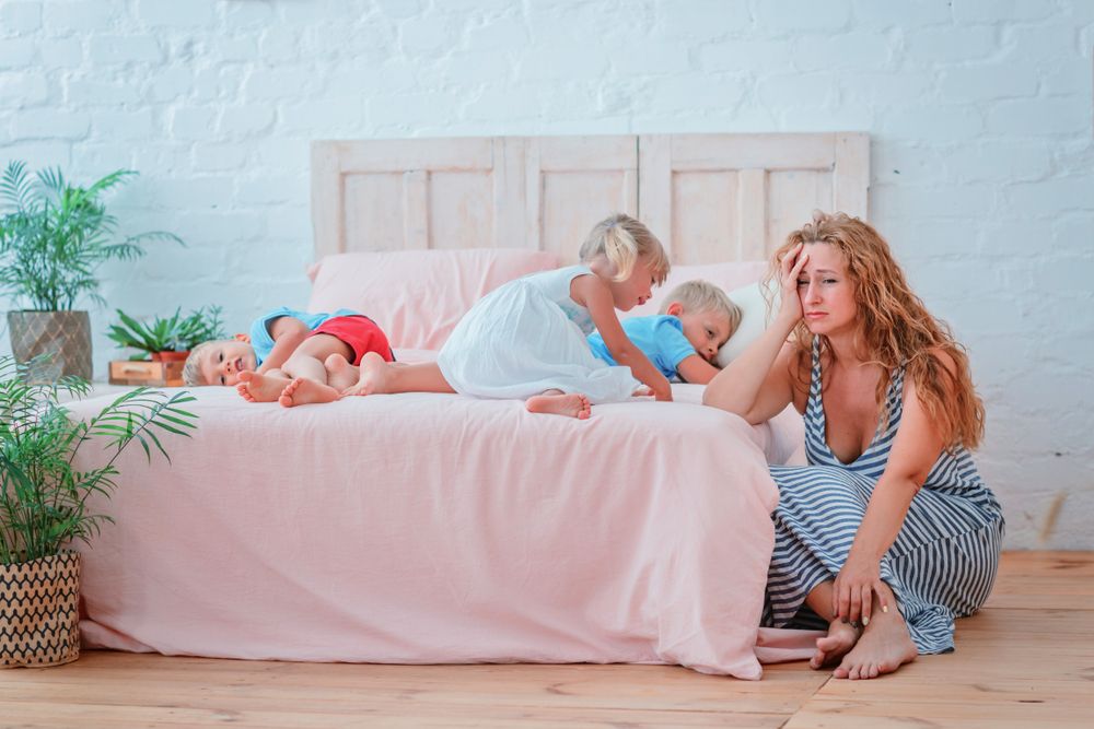 Tired mother with many children sitting near a bed with disobedient children