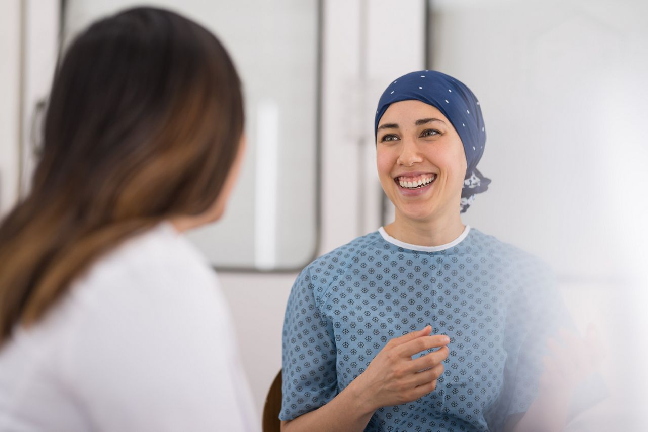 A millennial-age ethnic woman battling cancer talks with her doctor at the hospital. She is wearing a headcovering and smiling. The shot is over the doctor's right shoulder.