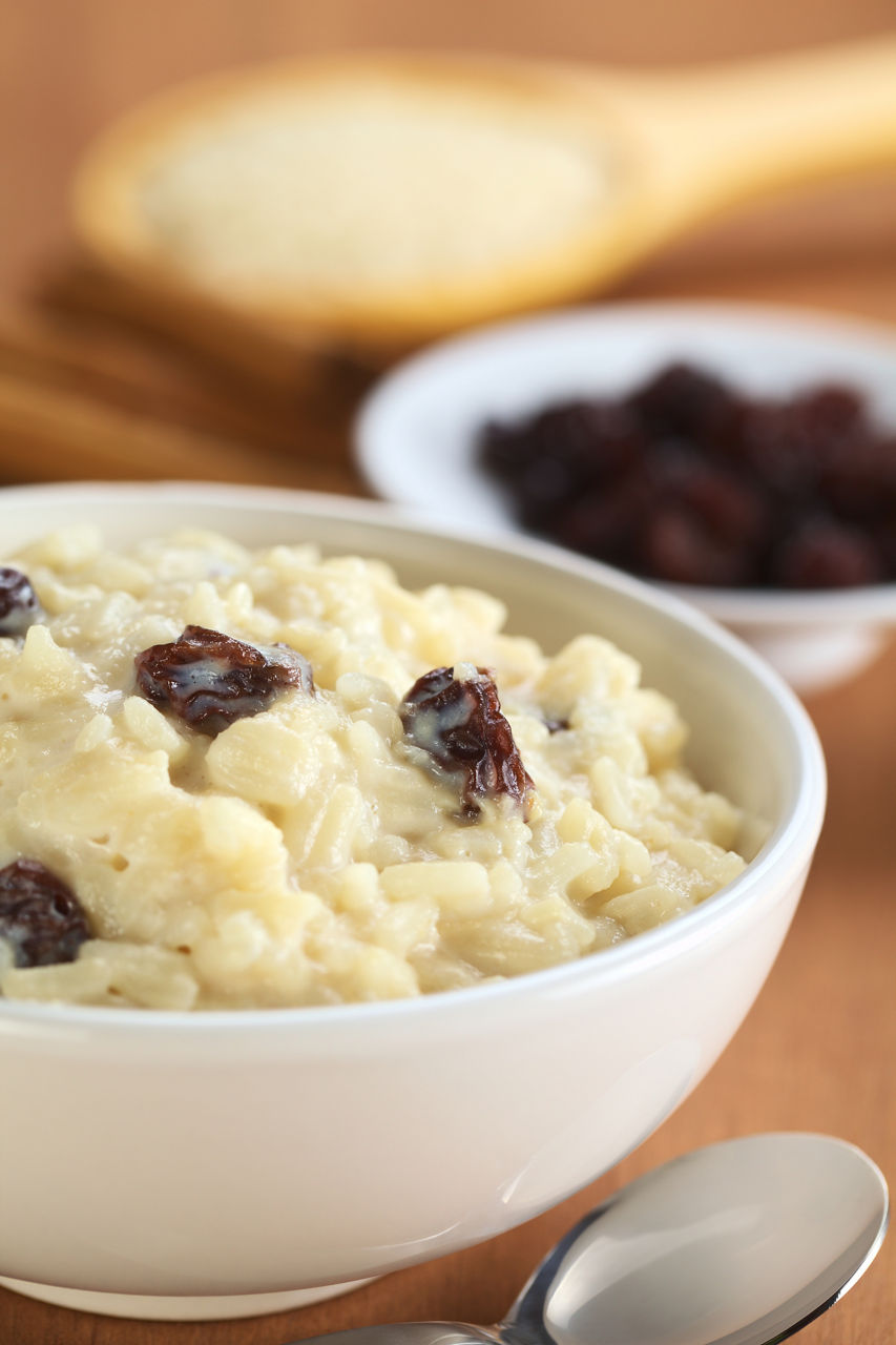 Delicious homemade rice pudding with raisins; with raisins, cinnamon sticks and raw rice on wooden spoon in the back (Selective Focus, Focus on the two raisins in the middle of the bowl)