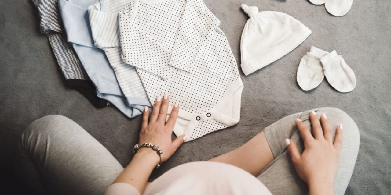 Une femme enceinte est assise sur le sol avec des vetements de bebe plies, notamment une grenouillere a pois, un chapeau et des mitaines. Le cadre est cosy et neutre, avec un eclairage doux mettant en valeur les textures delicates des vetements. La scene transmet la preparation et l anticipation d un nouveau-ne.