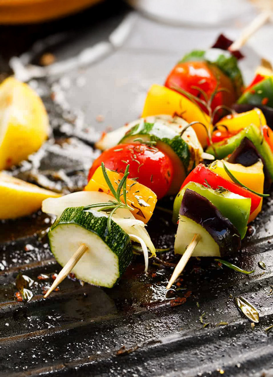 Colorful vegetable skewers featuring zucchini, bell peppers, tomatoes, and onions are arranged on a grill surface. The setting highlights a freshly cooked meal with visible herbs and spices. Lemon wedges are placed nearby, adding a vibrant touch to the composition.