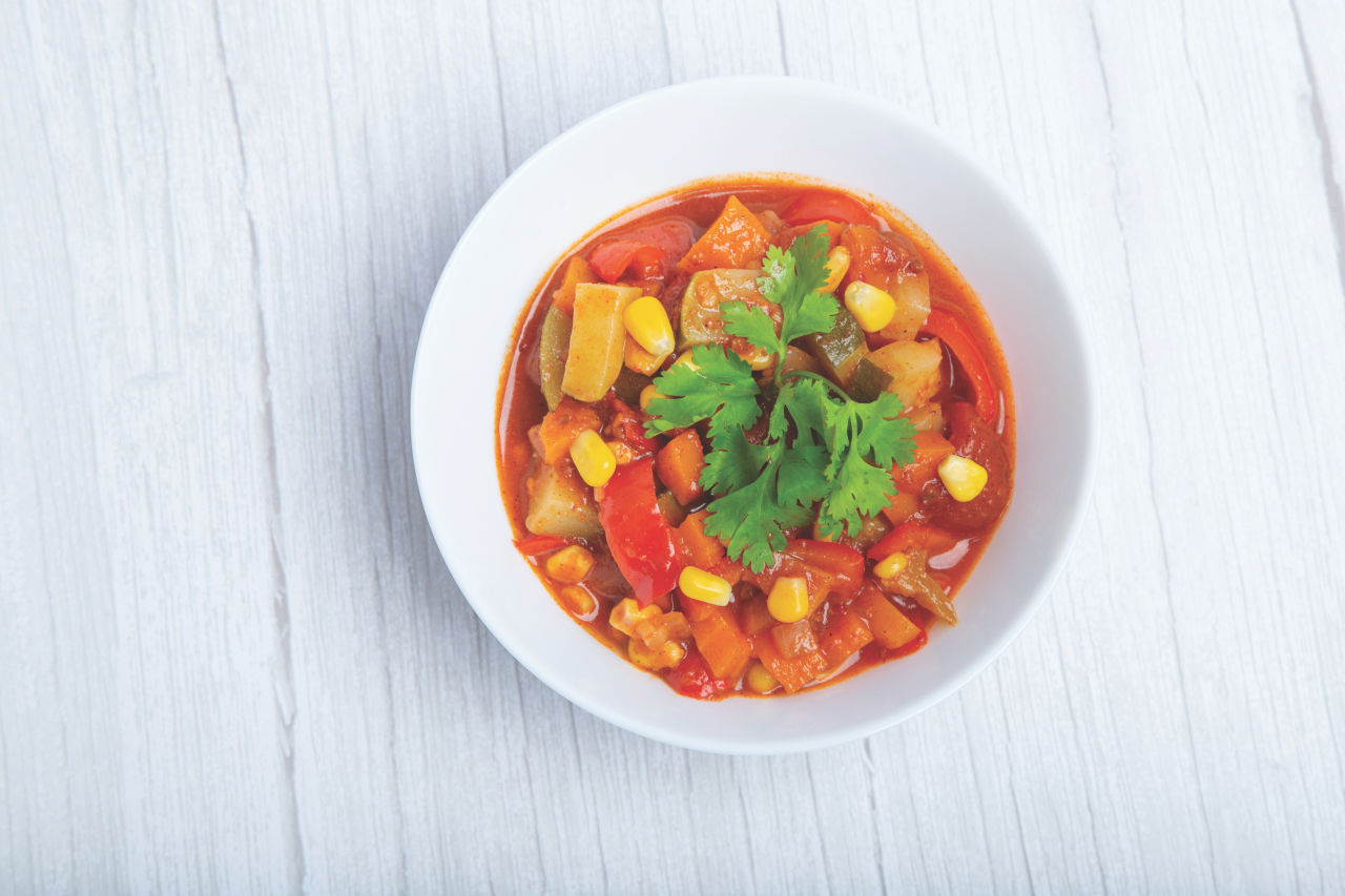 A vibrant vegetable stew served in a white bowl, featuring colorful ingredients like corn, tomatoes, and zucchini. The dish is garnished with fresh cilantro leaves, adding a touch of green to the presentation. The bowl is placed on a textured white wooden surface, enhancing the rustic aesthetic.