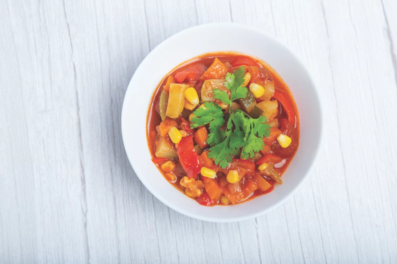 A vibrant vegetable stew served in a white bowl, featuring colorful ingredients like corn, tomatoes, and zucchini. The dish is garnished with fresh cilantro leaves, adding a touch of green to the presentation. The bowl is placed on a textured white wooden surface, enhancing the rustic aesthetic.