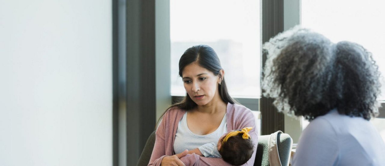 Eine Frau hält ein Baby im Arm und spricht mit einer zweiten Frau in einem hellen Raum mit großen Fenstern.