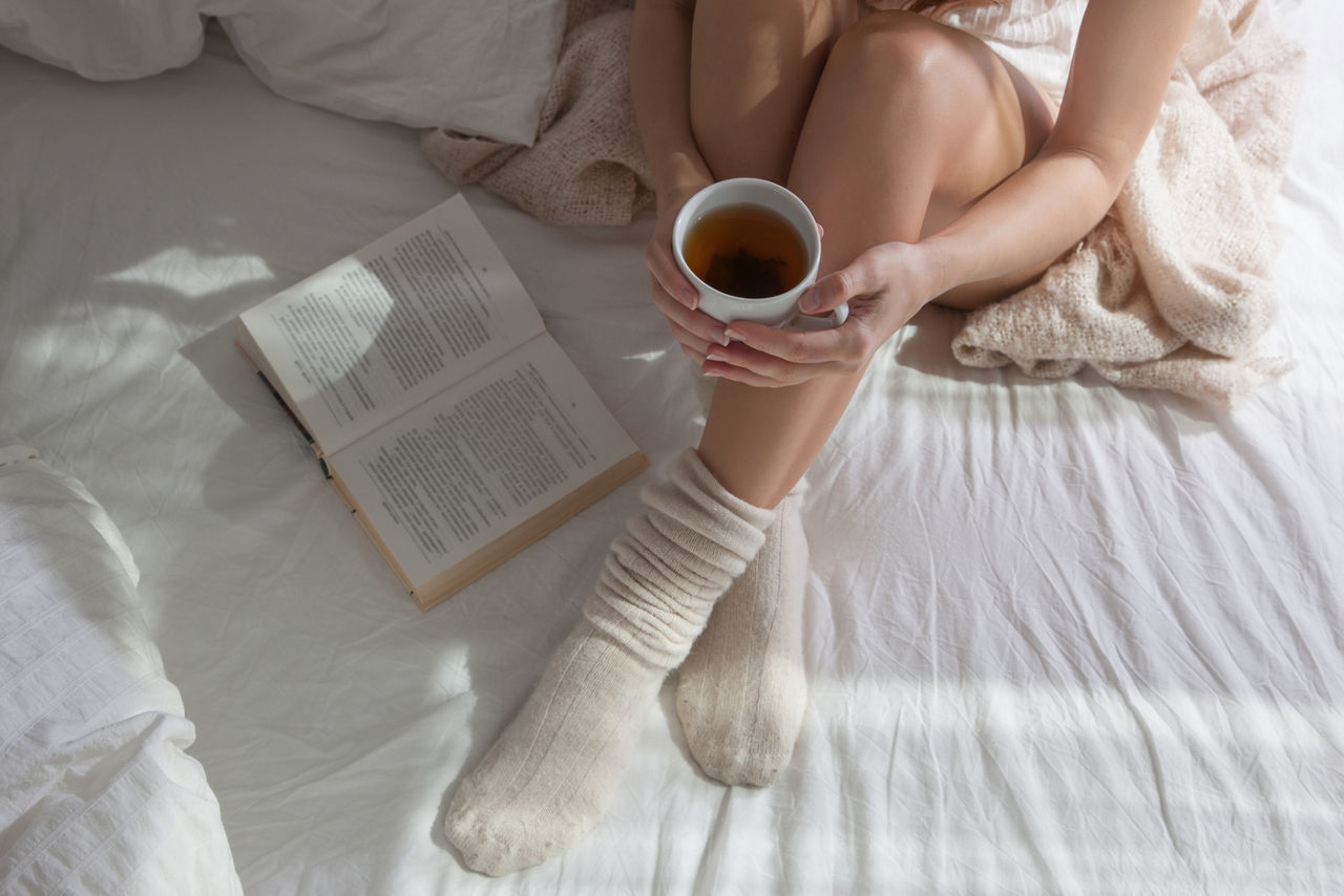 Long female feet in warm woolen socks around her feet is a book she is reading it.Part of the body.Beautiful female legs sitting on the bed with a cup in his hands.Morning with a book in their hands.; Shutterstock ID 329565530,woman sitting on bed in socks with tea and book