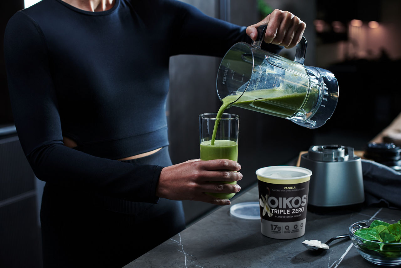 Woman pours green smoothie into glass, Oikos Triple Zero Vanilla yogurt on counter