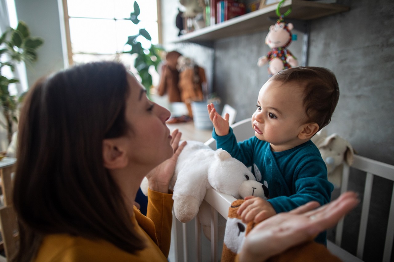 Cute baby boy in pajamas trying to explain something to his mother while standing in crib early in the morning